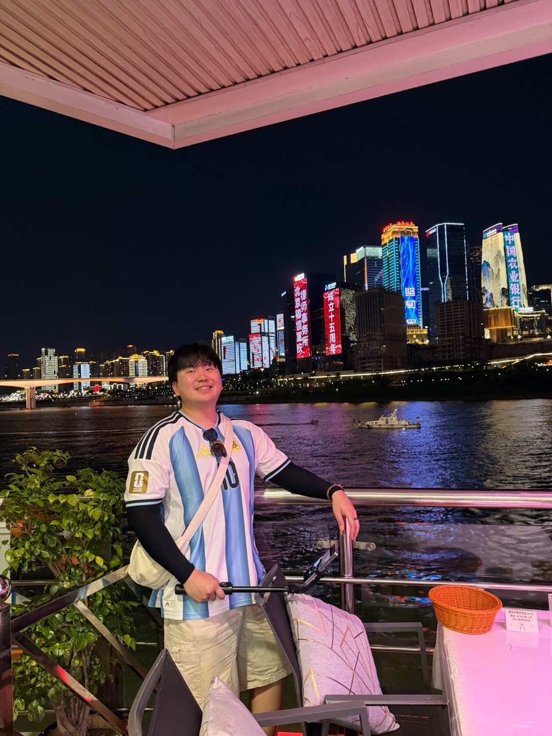 Junsu Kim smiling by the river with a city skyline at night
